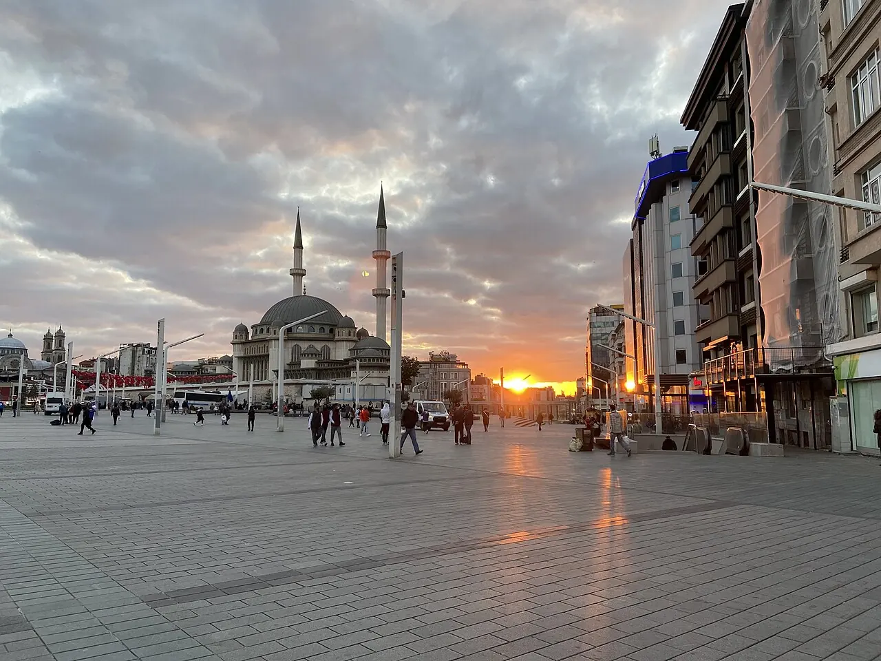 Pedestrians crossing Taksim Square beside the mosque building at sunset