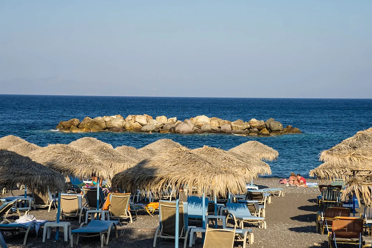 Dark volcanic beach, parasols and clear sea along the shore at Kamari